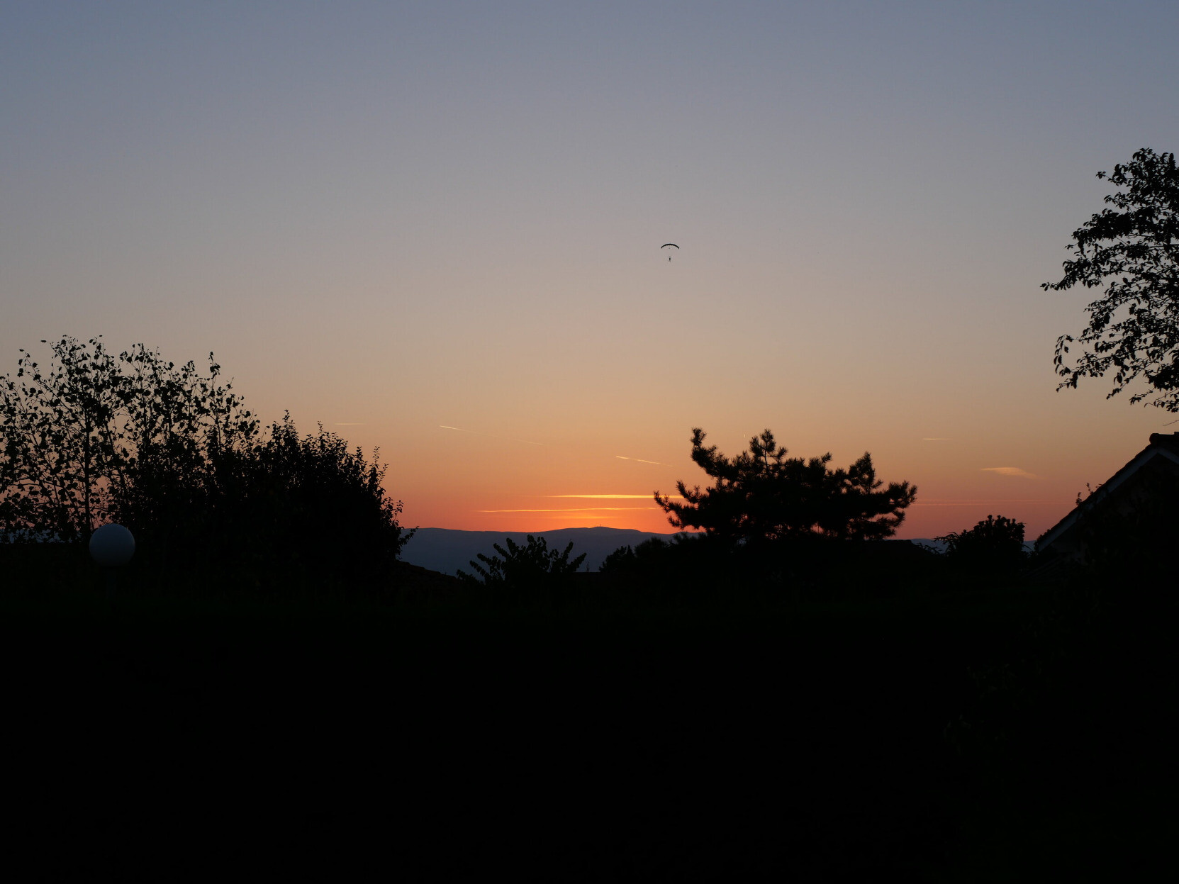 Couché de soleil orange derrière une montagne, un parachute se détache dans le ciel, ainsi qui plusieurs formes d'arbre et de maisons noires, dans le premier plan.