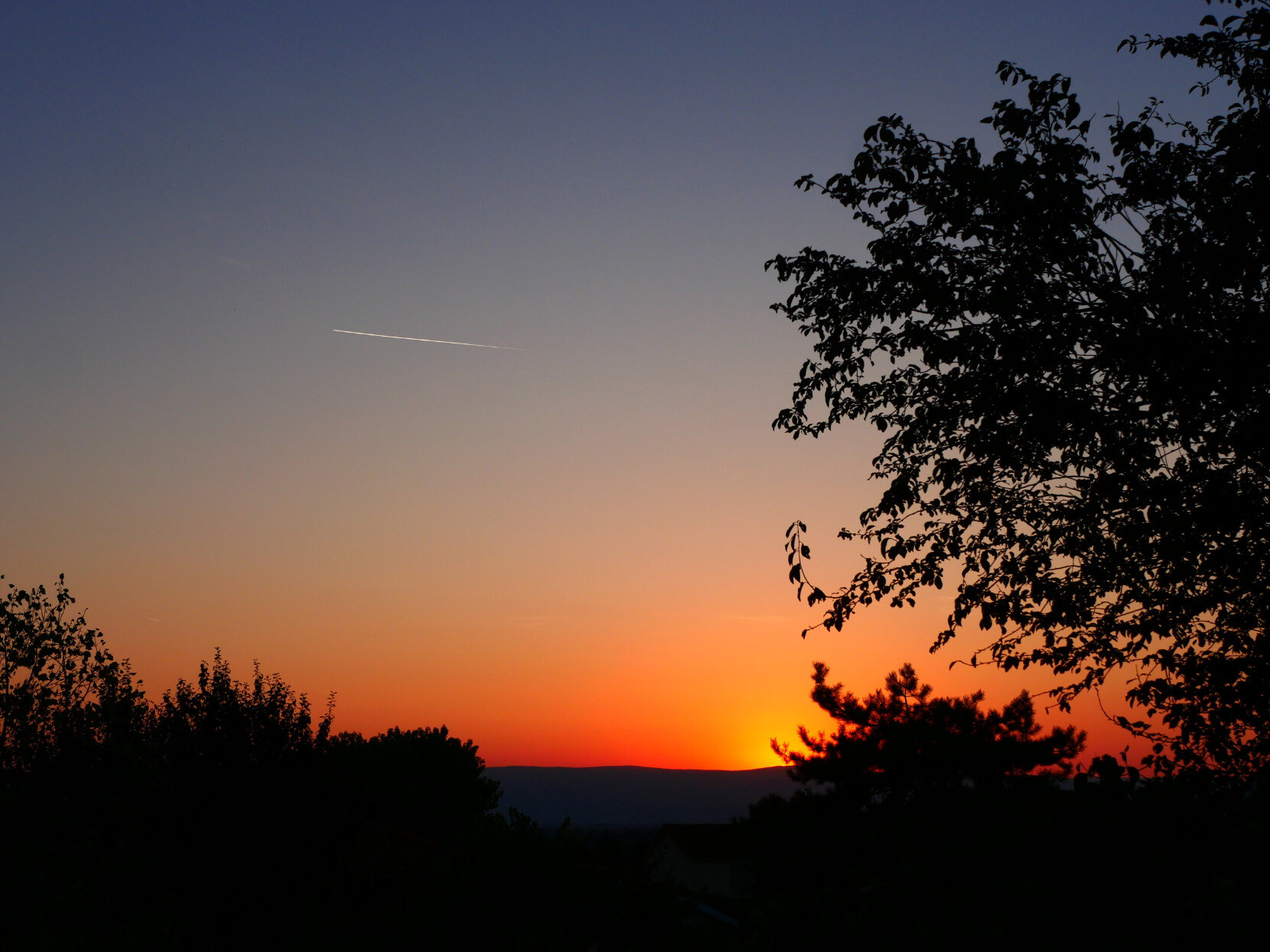 Couché de soleil, très orange derrière une montagne. La trainée blanche d'un avion encore éclairée par le soleil se détache dans le ciel qui s'assombrit. Des formes d'arbres noires se détachent en premier plan.