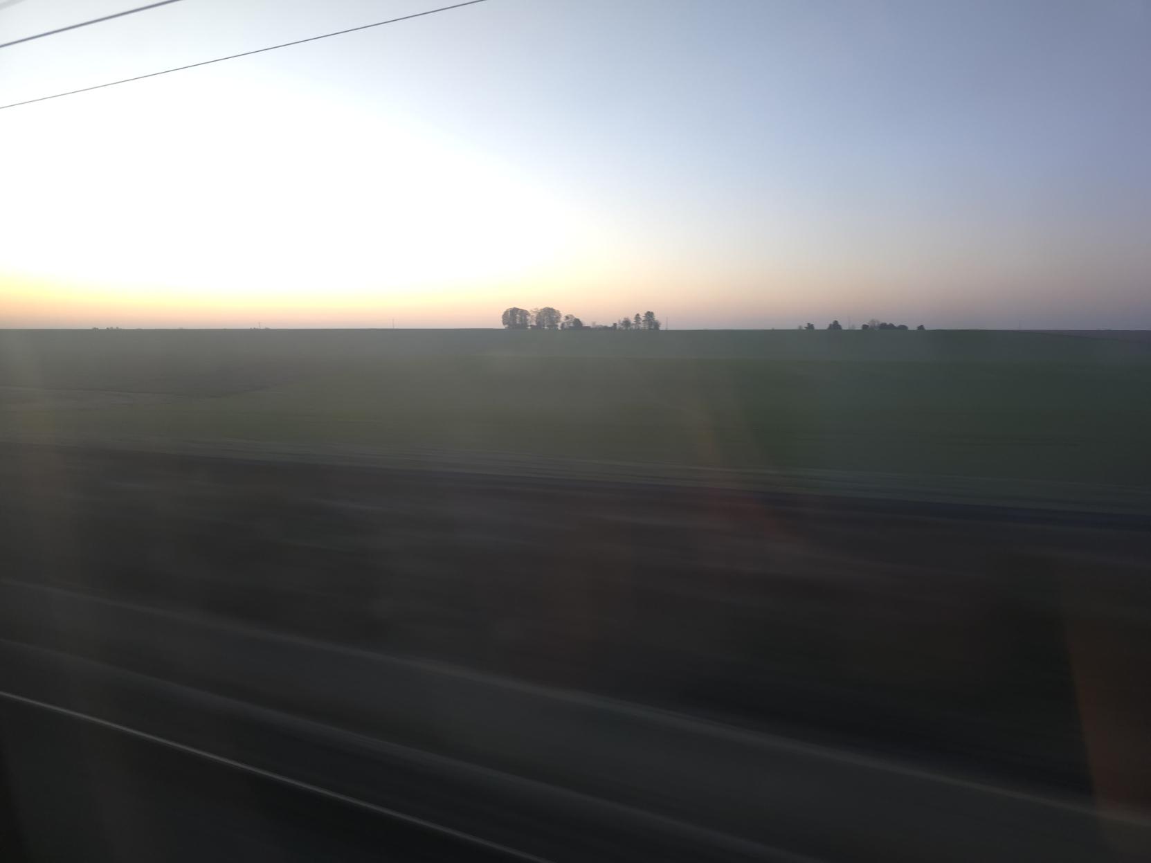 A photo of the sunrise seen through the TGV window. A small patch of trees stands in the distance between a dark green field and a hazy sky going from a bright orange to light blue. At the forefront, the blurry train track reminds us that TGVs go F  A     S       T.