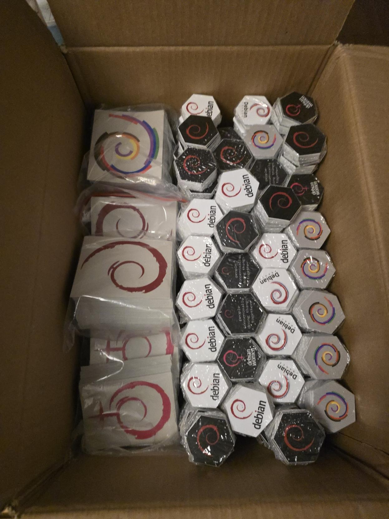 a photo of the inside of a cardboard box full of stacks of standard hexagonal stickers in various Debian designs, and some vinyl Debian, Debian Women and Debian Diversity swirls